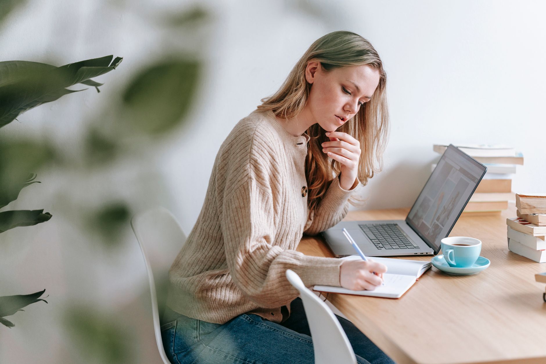 Woman writing in a notebook beside an open laptop and a cup of coffee at a wooden desk