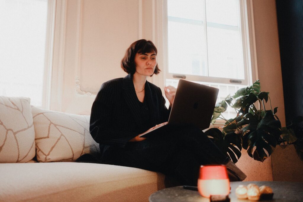 Woman in a dark pinstripe blazer working on a laptop on a sofa beside a bright window