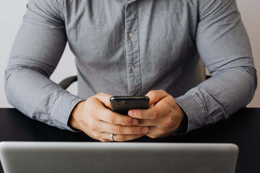 Person in a grey shirt holding a smartphone in both hands at a desk behind an open laptop