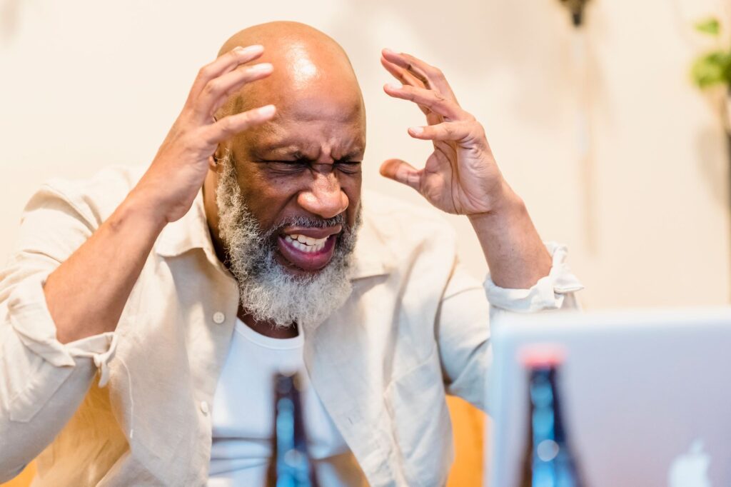Man with a grey beard raising both hands in frustration in front of an open laptop