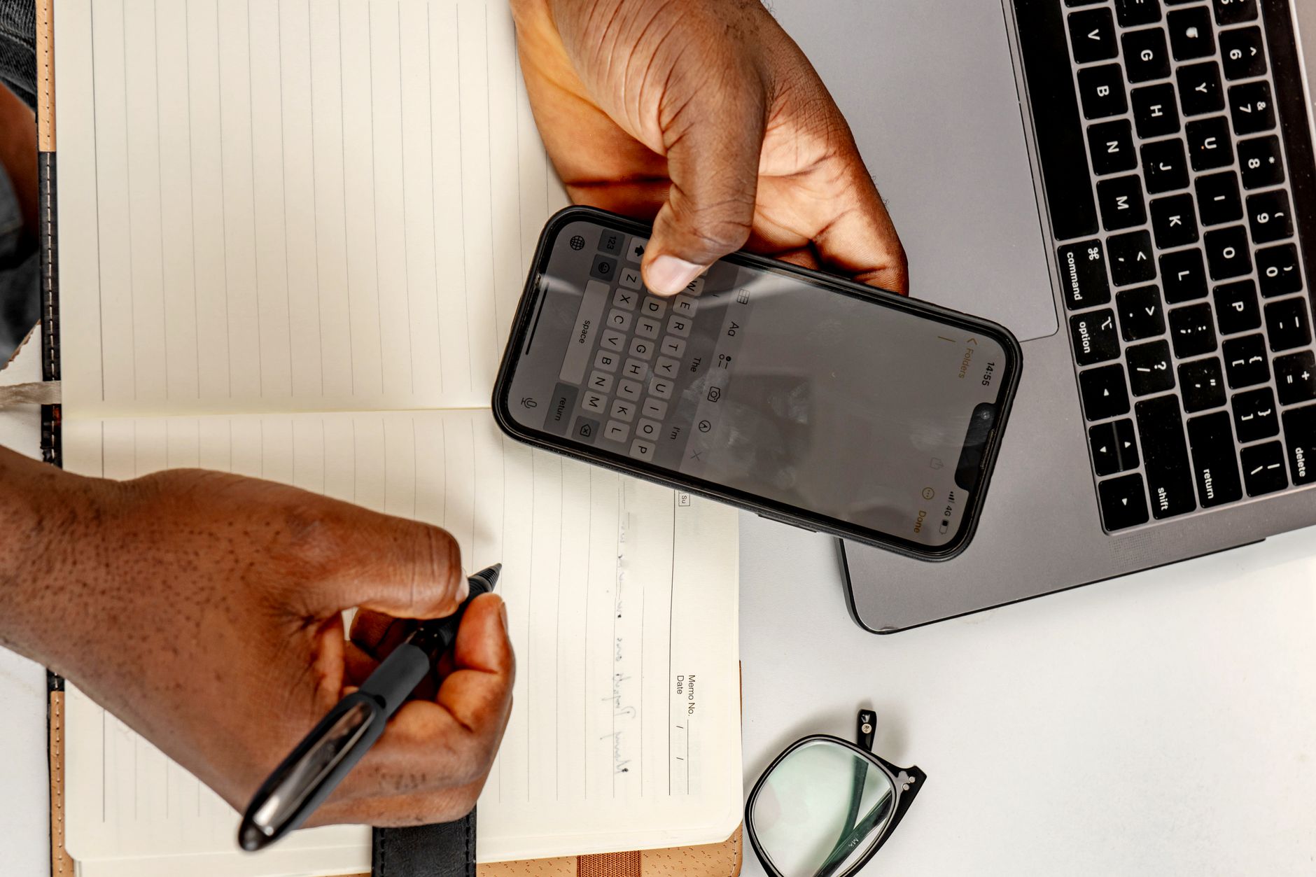 Overhead view of hands holding a phone with the on-screen keyboard open, beside a notebook and a laptop